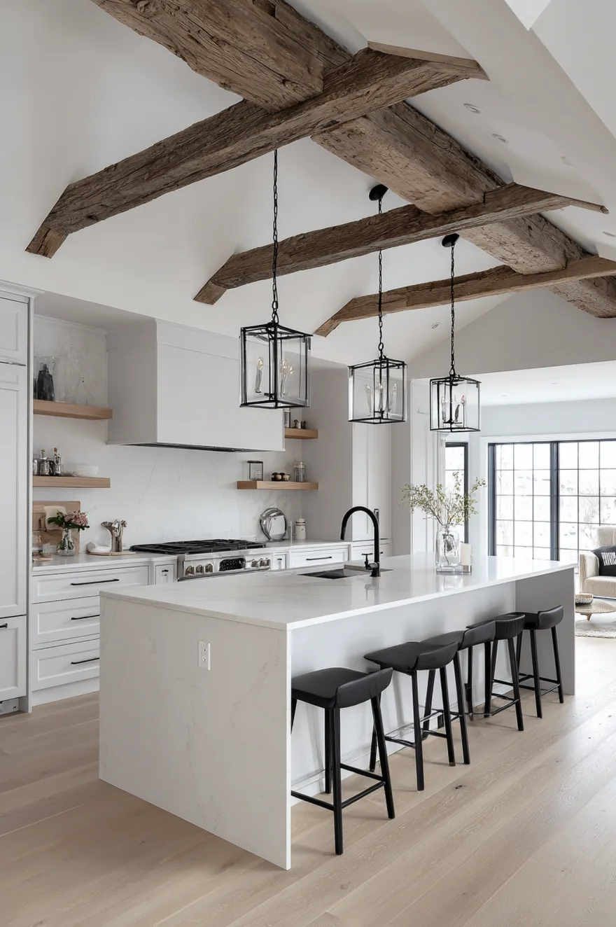 A white kitchen featuring a large island with black bar stools and three black metal lantern pendant lights. The vaulted ceiling is accented with thick rustic wood beams, highlighting the open concept design.