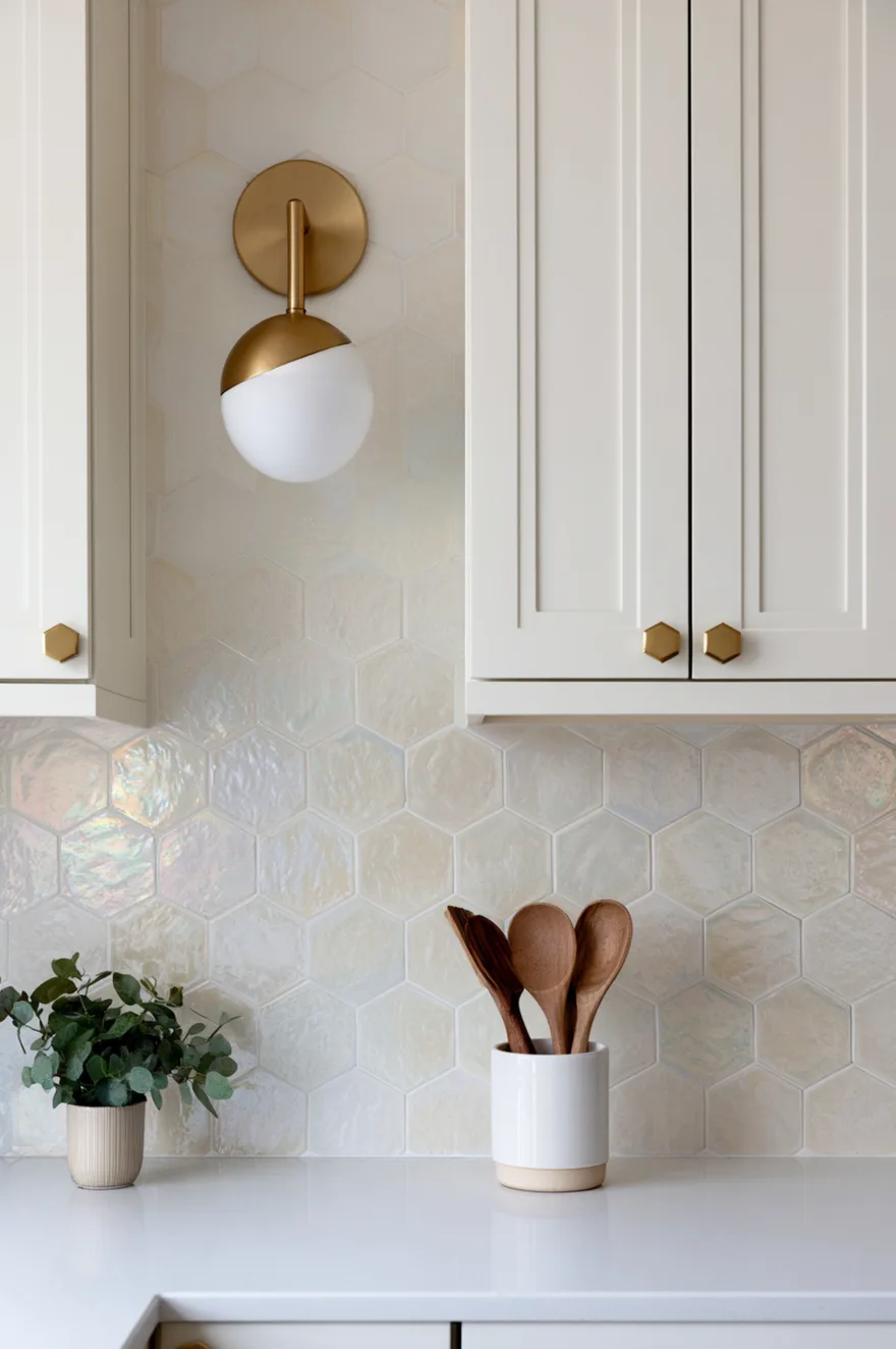 Large glossy white iridescent hexagonal honeycomb tile backsplash, white cabinets with simple brass hexagonal knobs, a modern round gold and glass orb wall sconce.