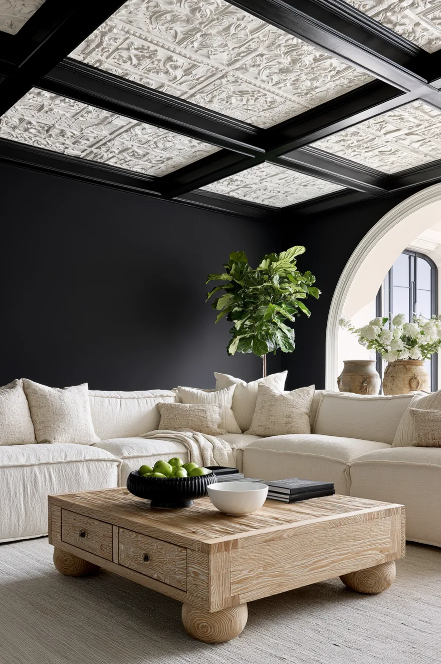 Cream linen sectional sofa and chunky wood coffee table against matte black wall with highly detailed ornate white ceiling.