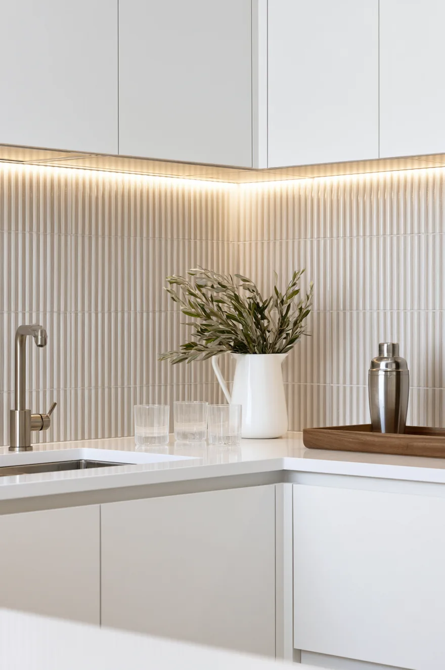 Off-white ribbed linear textured tile backsplash illuminated by warm LED strip lighting under white upper cabinets next to a white countertop.