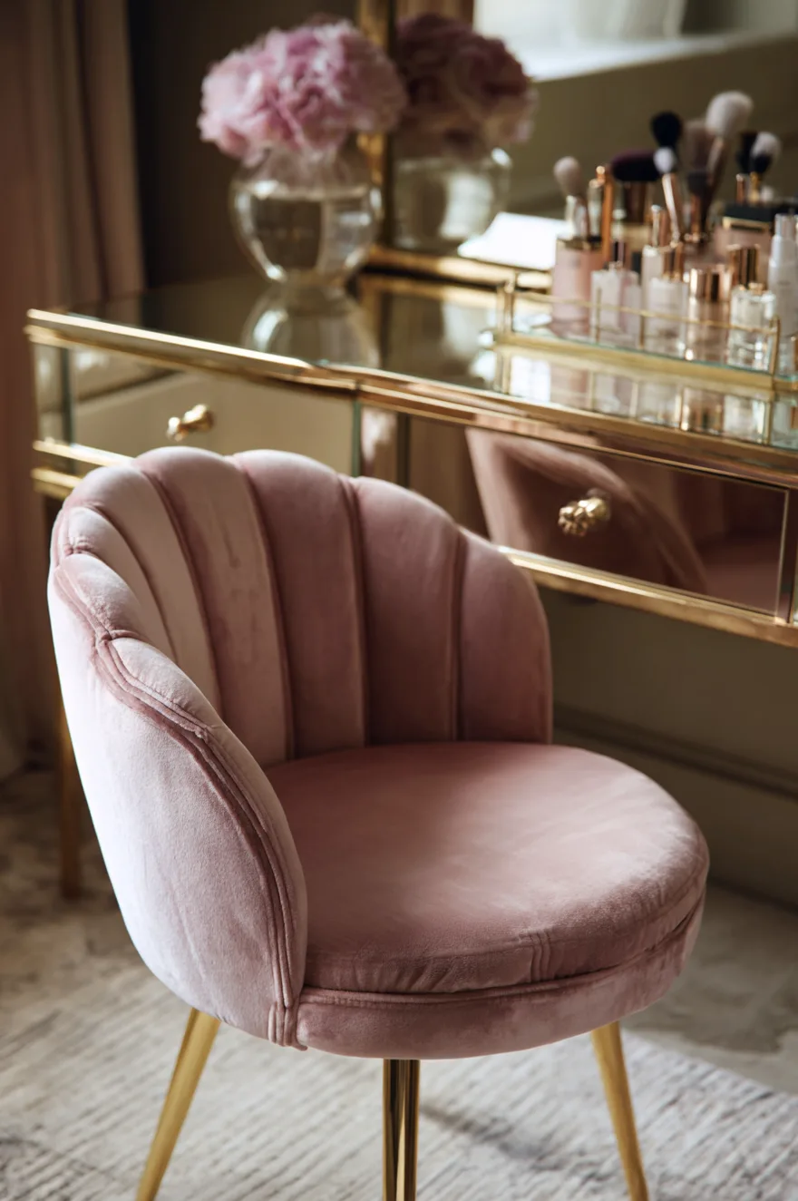 A close-up view of a pink velvet chair with a scalloped, shell-like backrest and gold metal legs. The chair sits in front of a highly reflective mirrored vanity desk with gold knobs. A gold tray holding makeup brushes and perfumes sits on top of the desk.